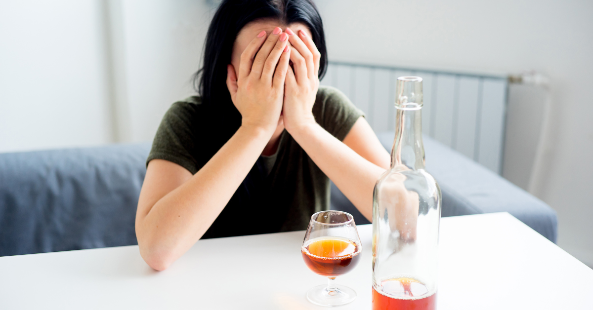 En person täcker för sitt ansikte med händerna. Bredvid finns en alkoholflaska och ett glas.
